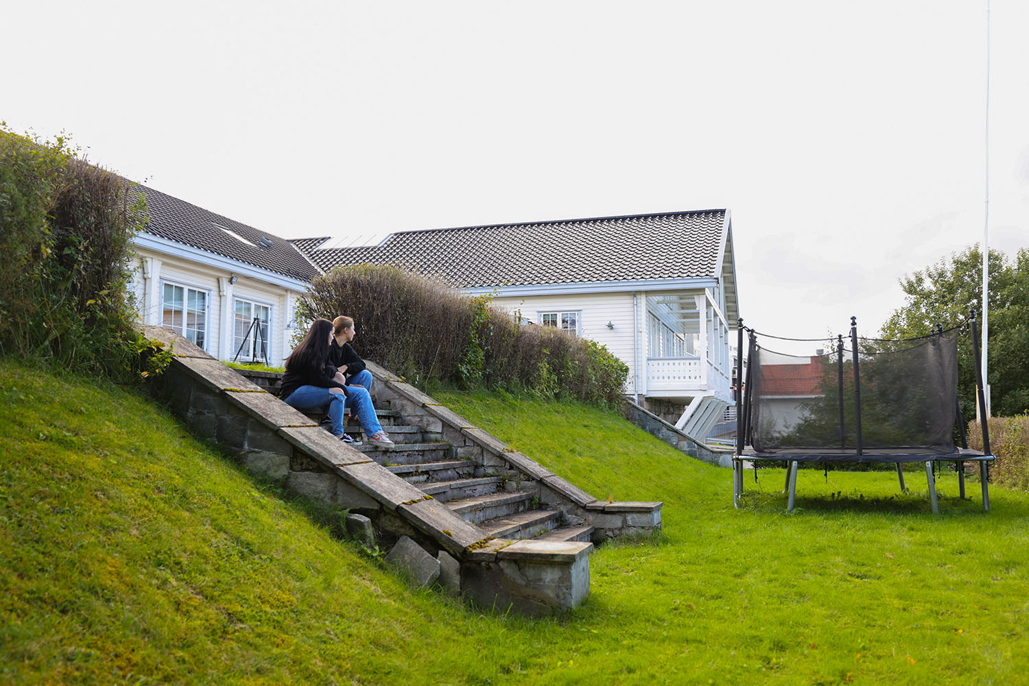 Hage med hekk, grønn plen, trampoline og stentrapp. Foto.